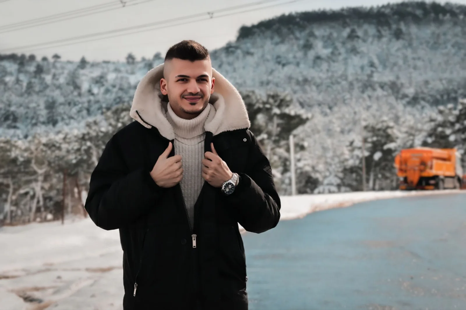Homme souriant en parka noire à capuche doublée sherpa dans un paysage enneigé