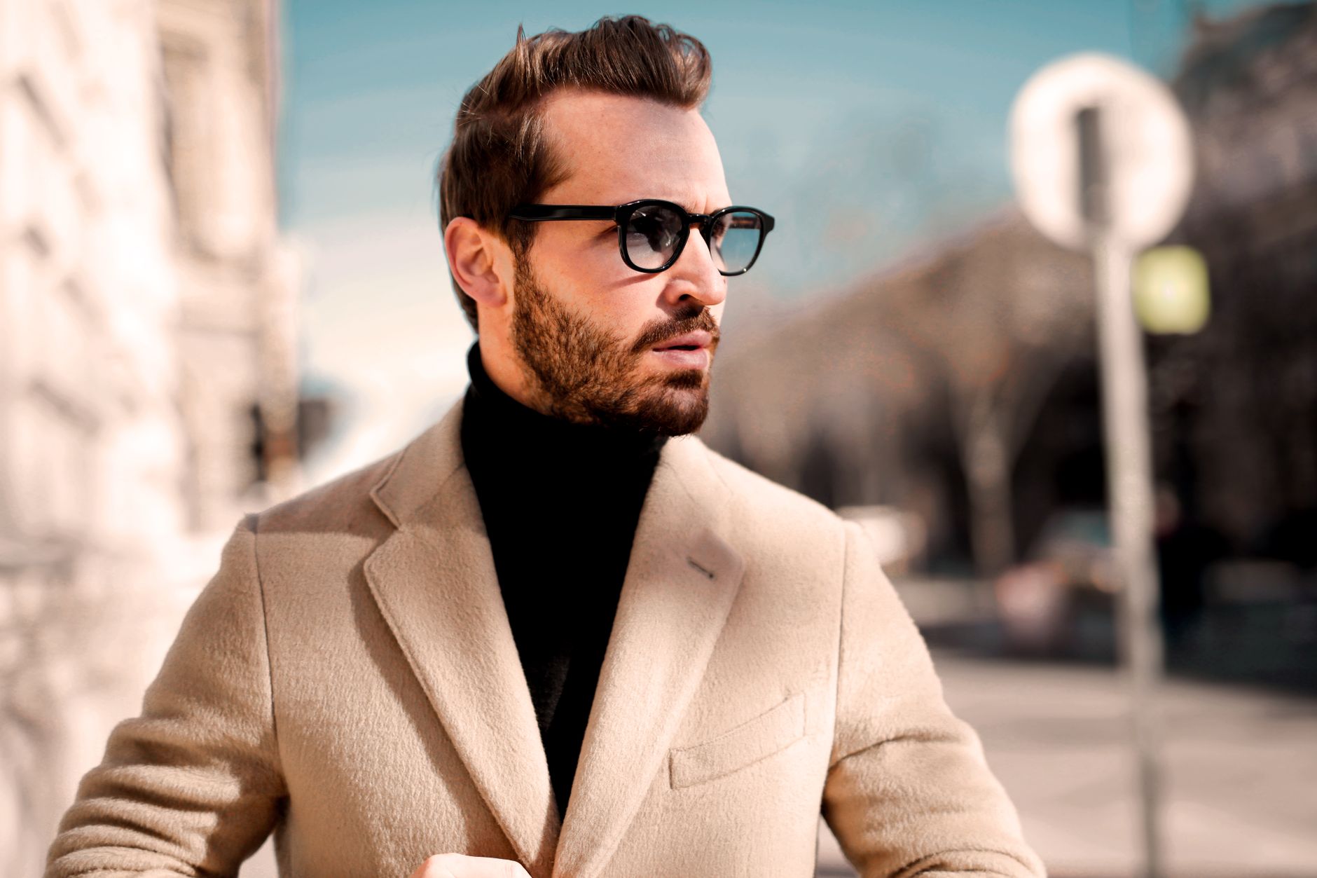 Homme élégant en manteau beige et col roulé noir dans une rue ensoleillée