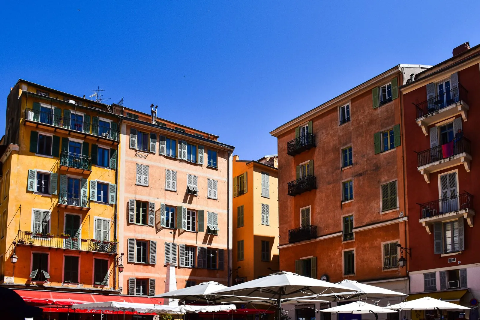 Façades ocre et orangées aux volets verts sur une place du Vieux Nice, parasols en terrasse
