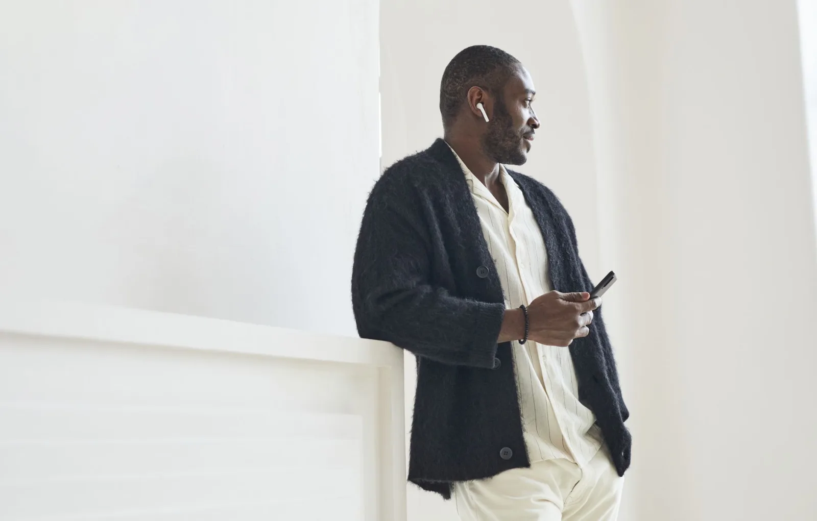 Homme en cardigan noir boutonné sur une chemise rayée, appuyé contre un mur blanc