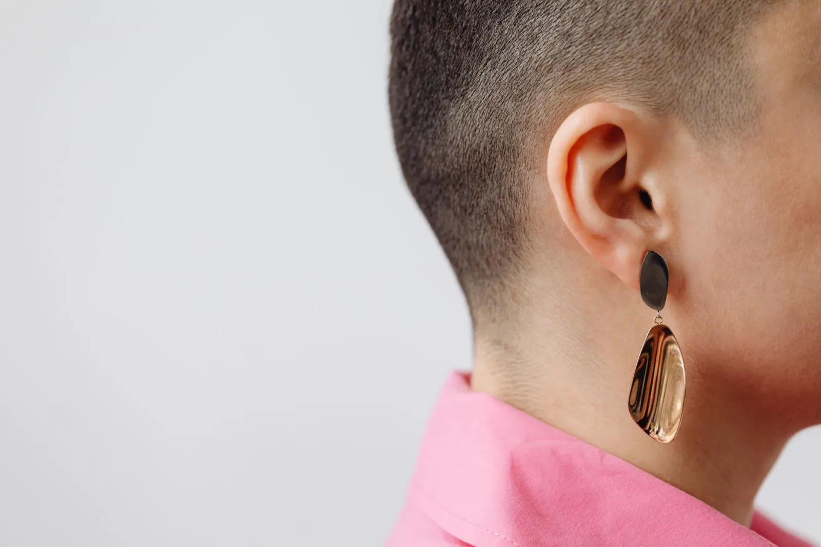 Ear of a woman with a shaved head wearing gold jewellery, on a light background